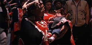 A Kwakwaka’wakw women in ceremonial regalia is cradling a baby who also is dressed in ceremonial regalia.