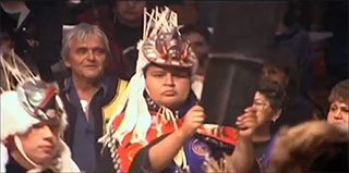 A young Kwakwaka’wakw man dressed in ceremonial regalia holding aloft a copper, behind him stands Chief Cranmer.