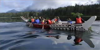 A large Kwakwaka’wakw canoe is moving diagonally across the screen over calm waters against a forested and mountainous background.