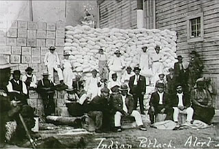 A group of Kwakwaka’wakw men are standing or seated in front of a large stack of flour sacks and boxes that have been gathered to present at a potlatch.