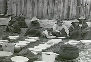Kwakwaka’wakw women in traditional dress sit on the ground next to food containers used in preparing food for a potlatch