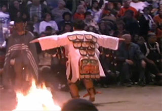 The back of a dancer wearing an embroidered tunic next to a fire pit in the big house.