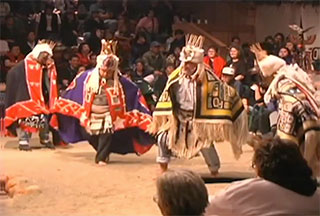 A group of dancers perform in woven ceremonial regalia to the right of a fire pit in the big house.