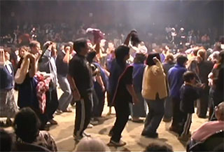 A group of people move in circles around the dirt floor at the center of the big house, they are surrounded by a large audience.