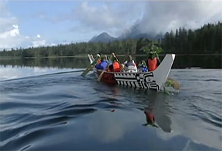 Image alt tag: A large Kwakwaka’wakw canoe is moving diagonally across the screen over calm waters against a forested and mountainous background.