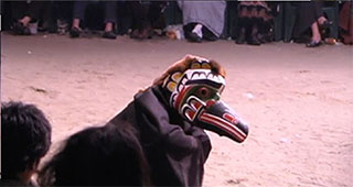 Un danseur de potlatch portant un masque aviaire et une cape noire s’accroupit et sautille le long du sol en terre de la maison cérémonielle.