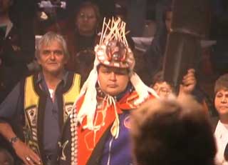 Image of a Kwakwaka’wakw dancer at a potlatch wearing ceremonial regalia consisting of a button blanket, carved and decorated frontlet.