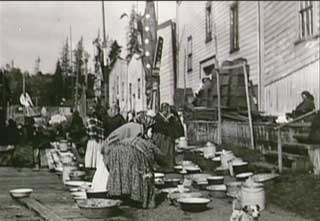 Historical black and white photograph showing a group of Kwakwaka’wakw adults standing among gifts gathered in preparation for a potlatch in Alert Bay.