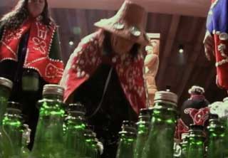 Image shows a figure wearing a woven cedar hat and button blanket regalia before a group of bottles which have been filled with oolichan oil.