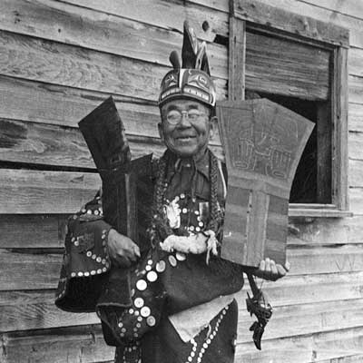 Chief Seaweed stands before a wood building, wearing a button blanket, ceremonial frontlet and holds a copper in each arm.