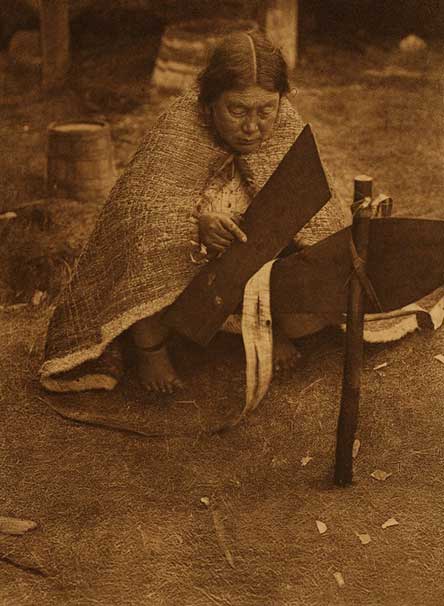 Elderly woman sits on cedar mat wearing a cedar bark blanket shown softening cedar bark using a tool called a bark beater.