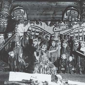 Three men in regalia dance around a fire, behind them a group of masked men stand before a painted screen and totem poles.