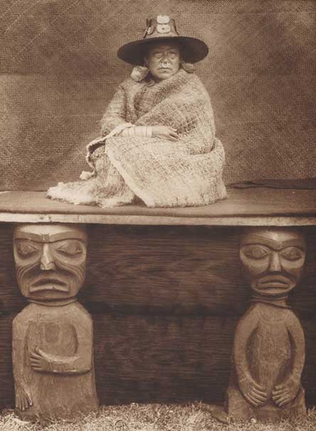 Femme assise sur une plate-forme supportée par deux personnages sculptés en bois. Elle porte un chapeau tressé, de grosses boucles d'oreille, un anneau nasal et un châle d'écorce.