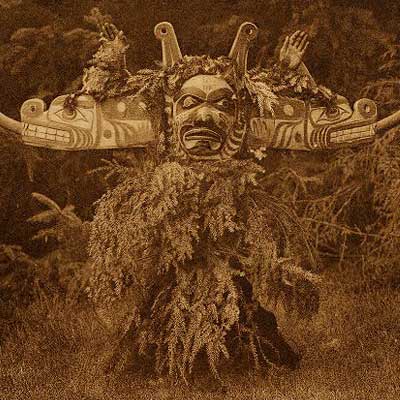 Sepia tone photograph of dancer in Sisiyutł mask with body covered in hemlock branches.