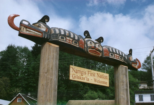 Large carved Sisiyutł or double headed serpent spans two upright posts with wooden sign bearing a welcome message.