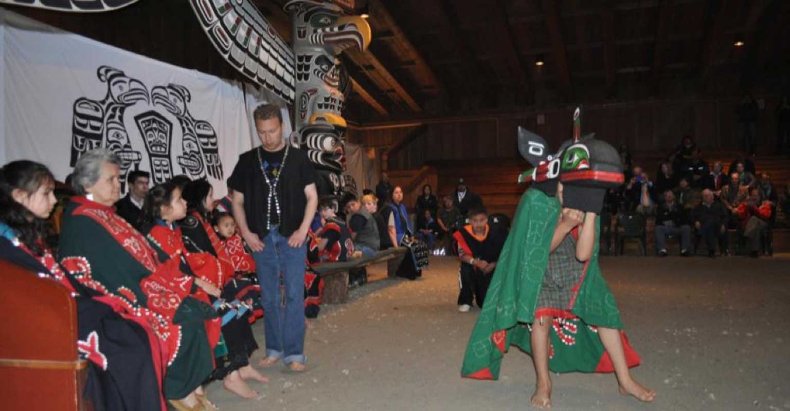Colour photograph of young Imas Dancer wearing a whale mask and green cape in front of gathered crowd in big house
