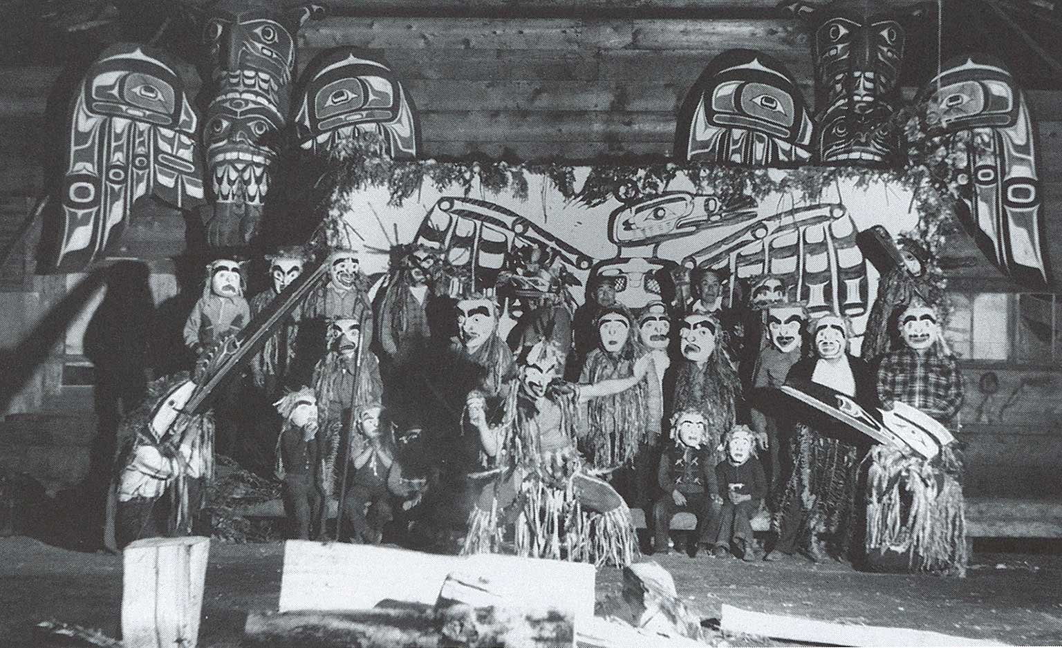Black and white photograph of a group of masked dancers in ceremonial regalia standing before a dance screen in the big house