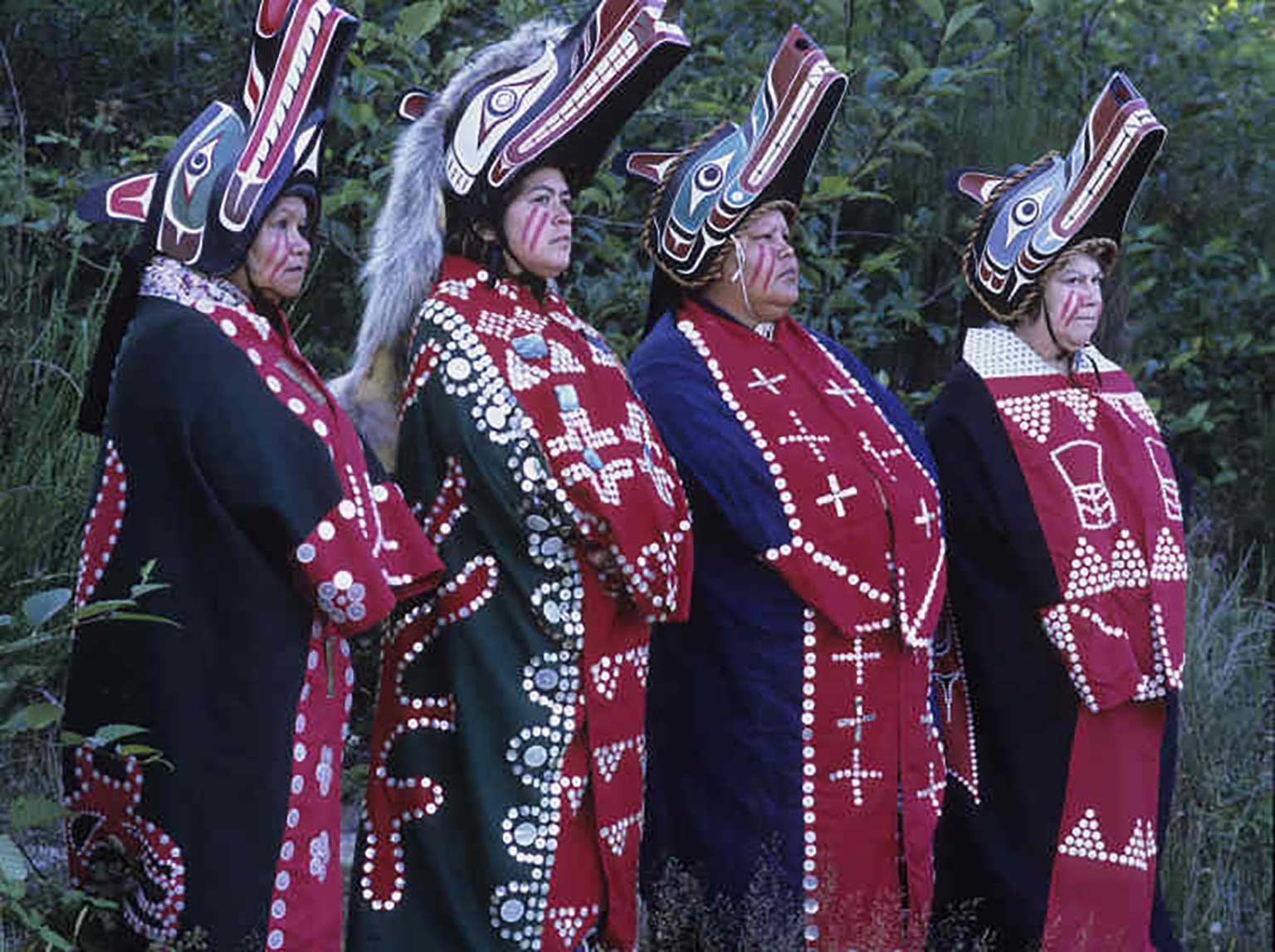 Colour photo of four women in a row facing to the right of the camera, each wears a button blanket and a wolf headdress.