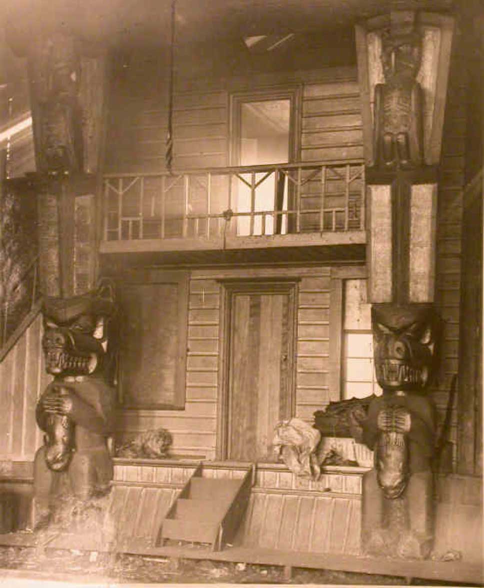 Photographie sépia de la façade d'une maison à deux étages avec porche, flanquée de deux hauts poteaux de maison en forme d'ours