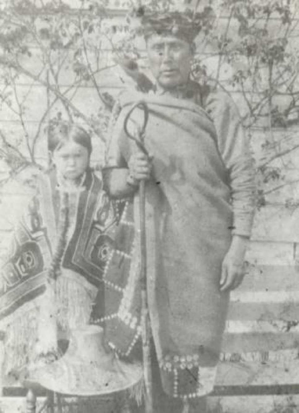 Photographie noir et blanc de Tsandigam Nege', Harry Mountain, portant un attirail d'écorce de cèdre, se tenant à côté un enfant vêtu d'une couverture à boutons