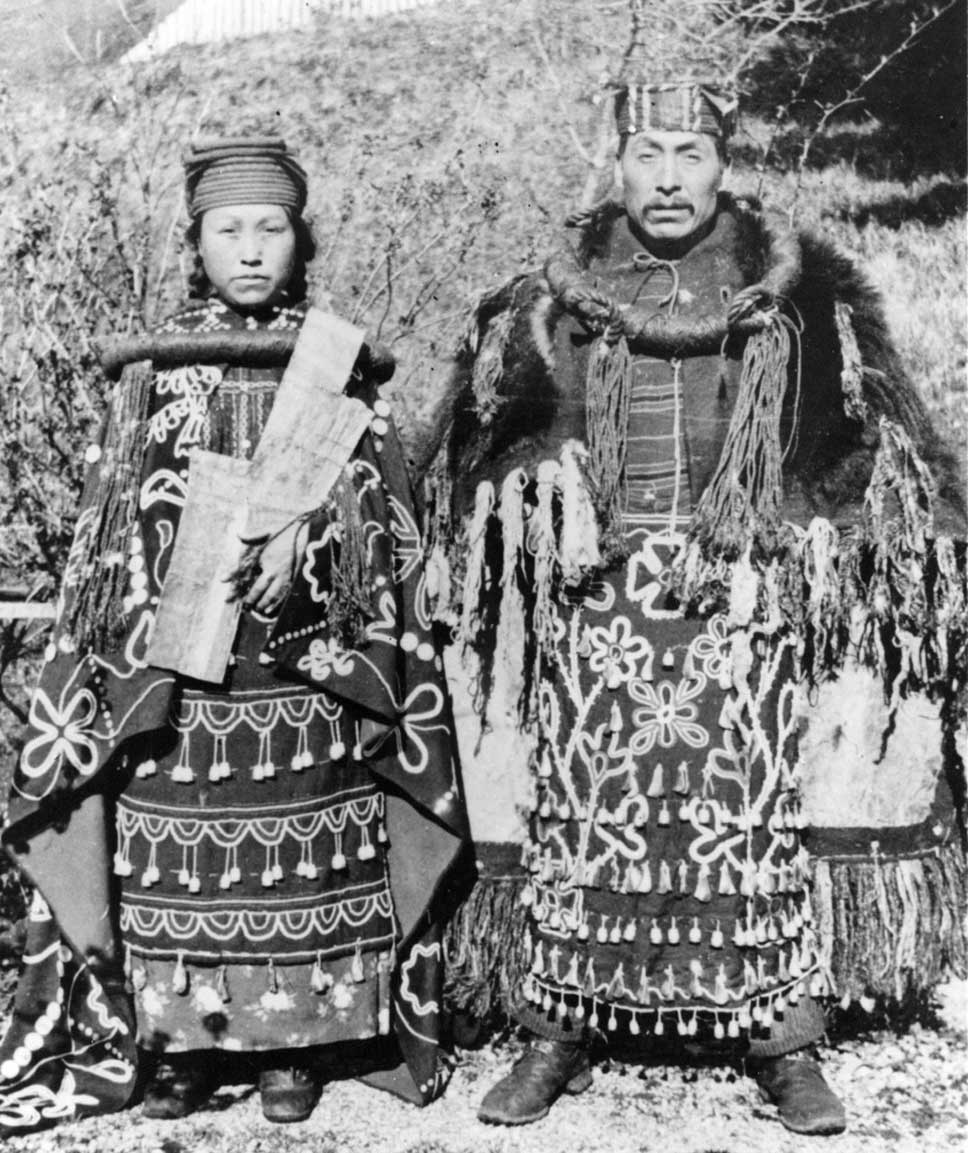 Black and white photo shows couple standing side by side in cedar bark regalia, dance aprons, button blankets, holding copper
