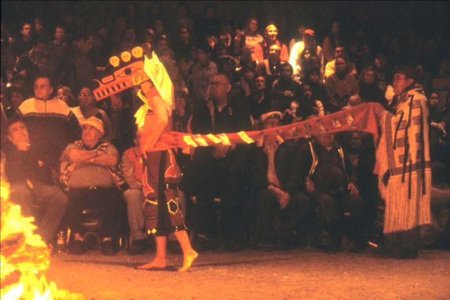 Danseuse aux pieds nus portant une couverture à boutons, suivi un ancien en tenue chilkat tenant une traînée, à la lueur du feu de la maison cérémonielle