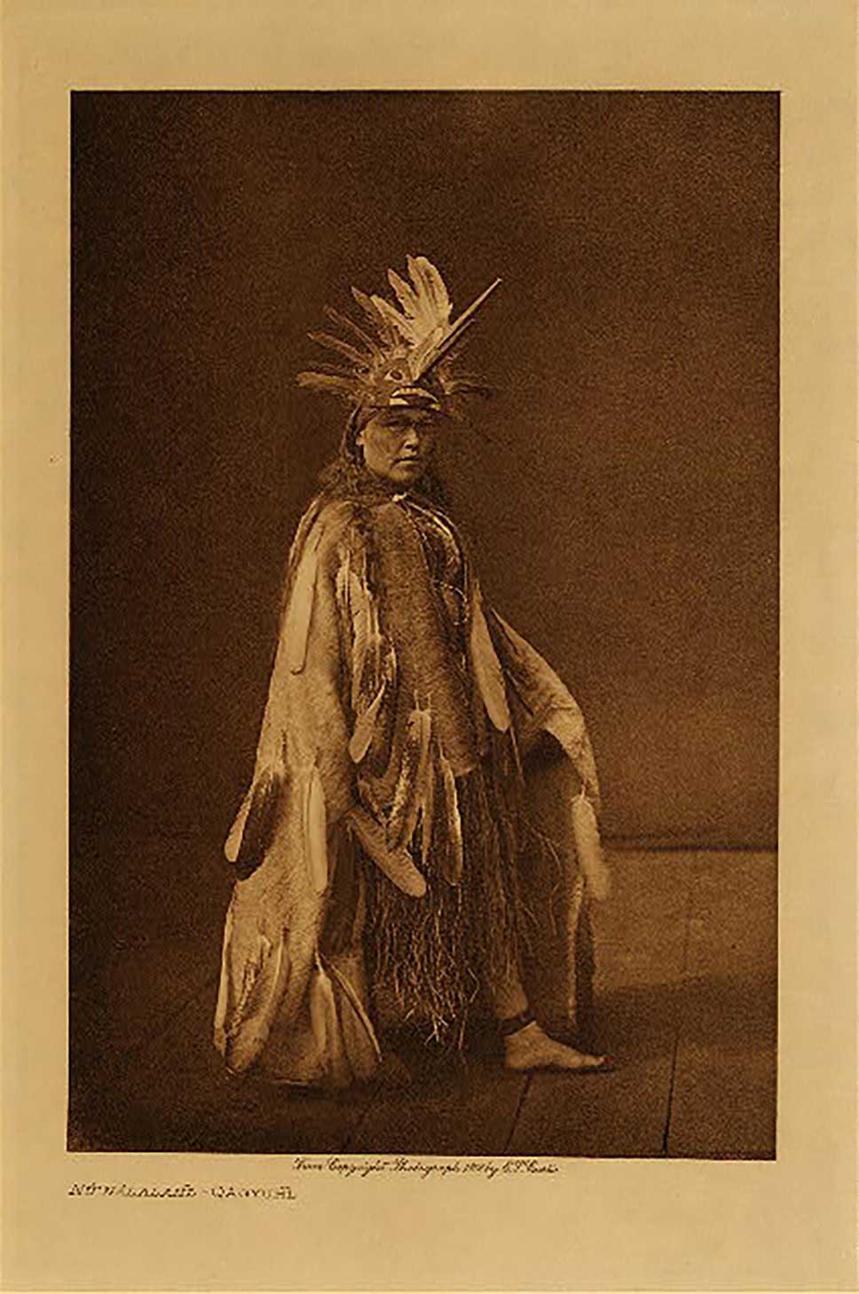 Photographie sépia par Edward Curtis, montrant un danseur en tenue cérémonielle portant une coiffe de la danse du Jour, 'Na'nalalał