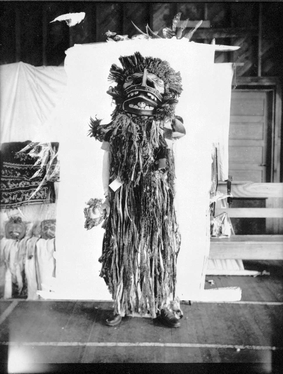 Homme posant devant un fond blanc, vêtu d'une coiffe à traînée d'hermine, tenant deux Cuivres, avec un masque d'aigle et un masque de loup à ses pieds