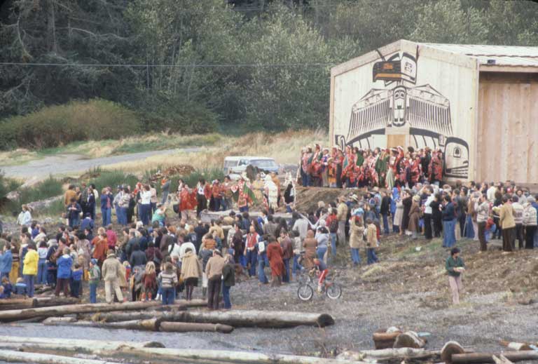 A large group of kwakwakw'wakw people, many in ceremonial regalia, assembled on beach and land in front of the U'mista Cultural Centre for its opening.
