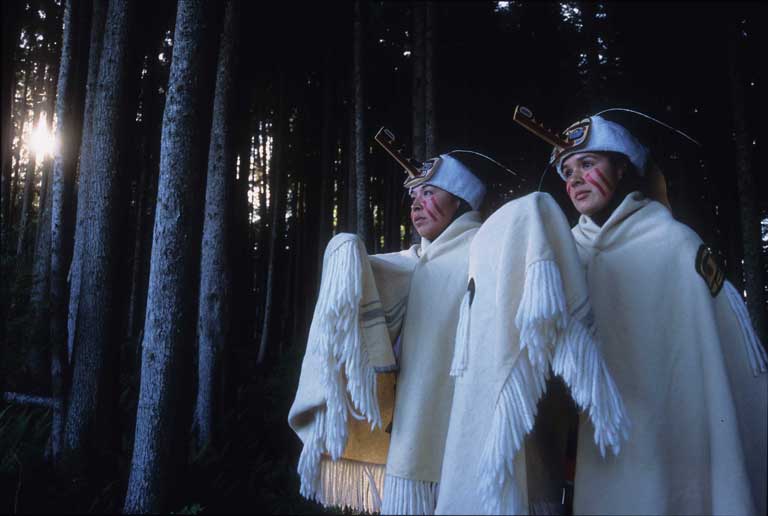 Two weather dancers in white wool and fur regalia, fur headdress and face paint, standing in a forest.