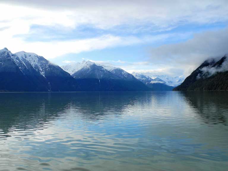 Calm ocean in foreground reflects a background of snow-covered mountains under a blue sky with scattered clouds.