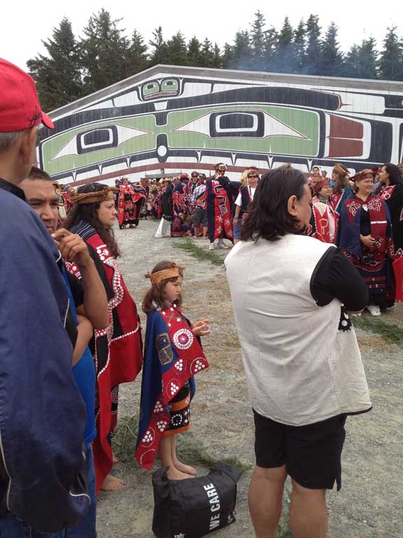 Kwakwaka'wakw men, women and children are standing in front of the big house, many wearing brightly cultured button blankets and other regalia.
