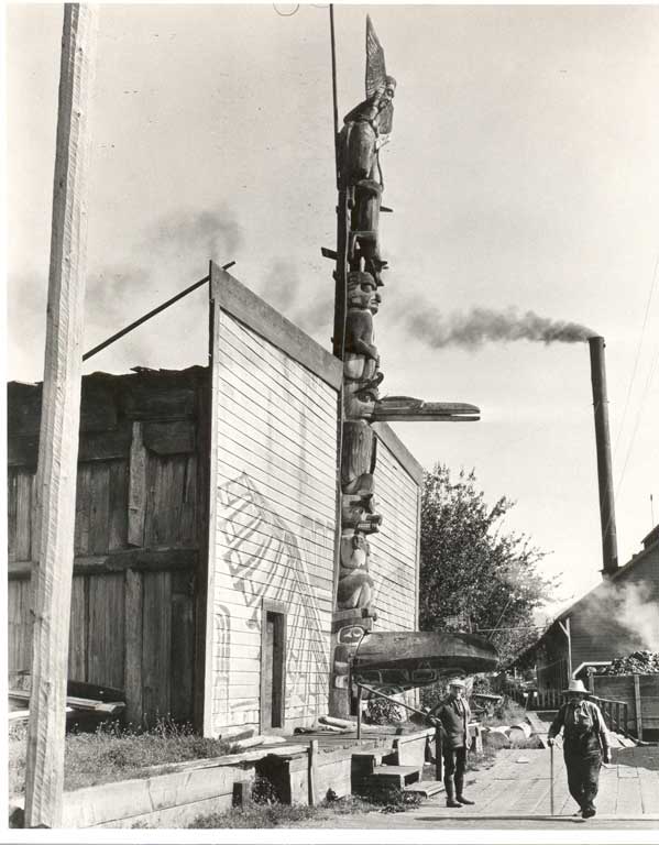 Deux hommes se tiennent devant une maison communautaire en bois à façade peinte et à grand mât central sculpté servant d'entrée.