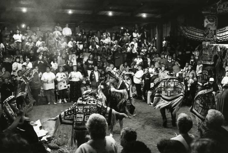 Three masked dancers wearing woven chilkat blankets dance before a group of spectators.