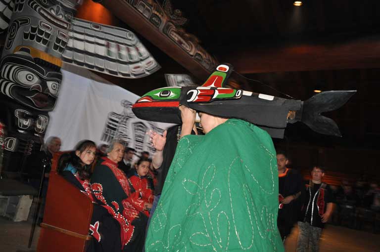 Colour photograph of dancer in whale mask, green robe, potlatch setting in big house, totem pole in background.