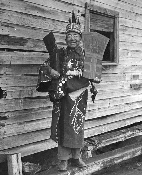 Chief Seaweed stands before a wood building, wearing a button blanket, ceremonial frontlet and holds a copper in each arm. 