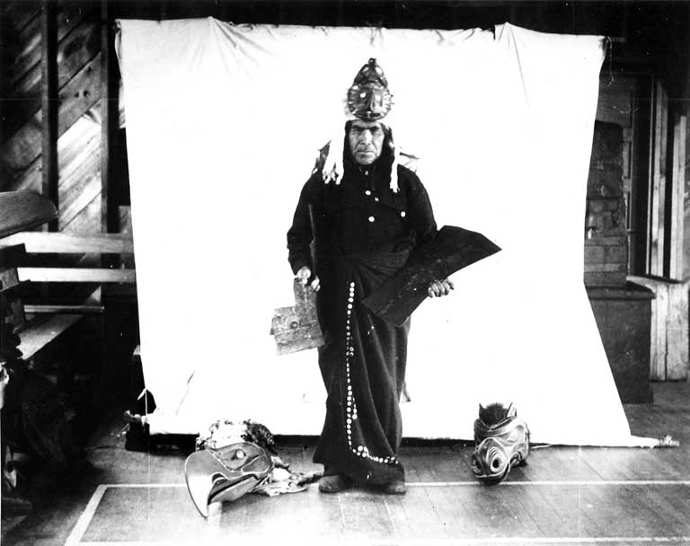 Un homme debout, vêtu d'une coiffe ornée d'hermine tient deux cuivres, un masque d'aigle et de loup à ses pieds, sur fond blanc.