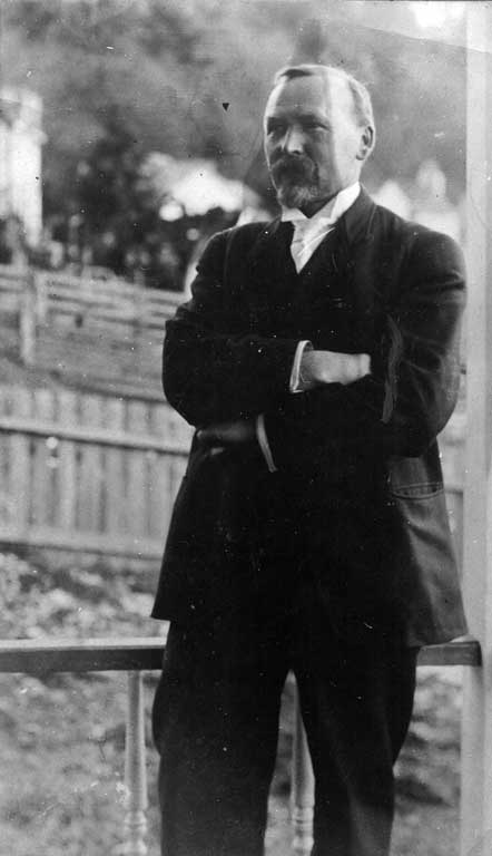 Un homme porte un costume noir à chemise blanche et cravate, les bras croisés, les cheveux courts et la barbe rasée de près.
