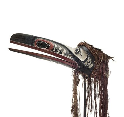 Un masque de corbeau au long bec articulé, peint en noir, lèvres rouges, narines sculptées, orbites et motifs en blanc, avec une frange d'écorce de cèdre.