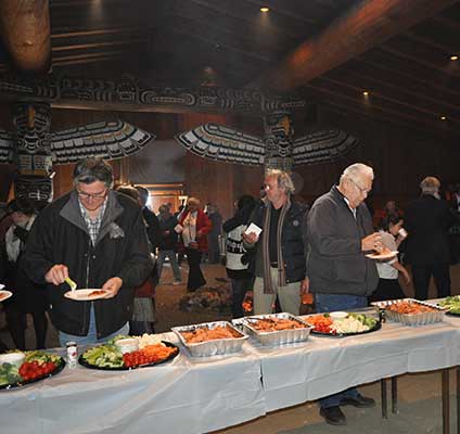 Photo couleur montrant une longue table de banquet, pleine de plats de saumon et de légumes, dans la maison cérémonielle lors un potlatch.