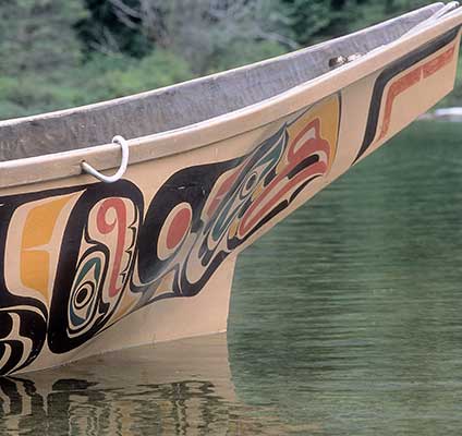 Colour photograph shows the bow section of a canoe, brightly painted figures adorn the hull.