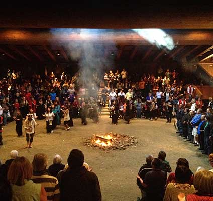 A large group of celebrants appear standing and dancing around a fire in the center of the bighouse.