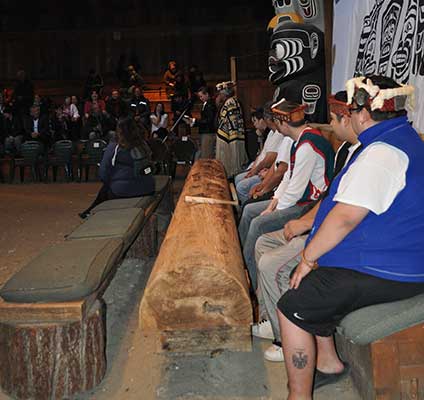Row of young men seated at a Tamidzu or drum log, behind them is a totem pole and dance screen.