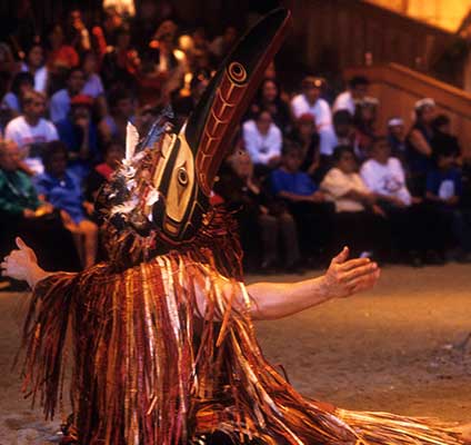 Colour photograph at potlatch in big house of dancer in regalia demonstrating the Tseka or red cedar bark ceremony.
