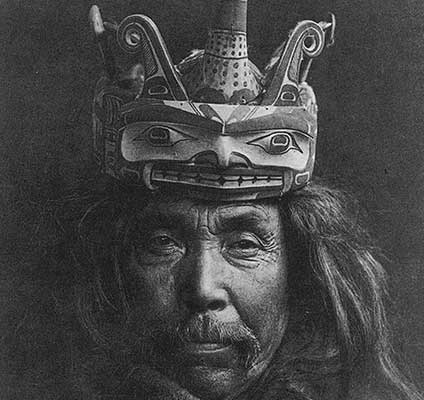 Black and white historic photograph of mature adult male, long hair and moustache wearing a carved headdress in the figure of a loon.