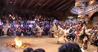 A group of dancers perform in woven ceremonial regalia to the right of a fire pit in the big house.