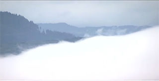 Un amoncellement de nuages blancs au premier plan, avec derrière un paysage de montagnes boisées.